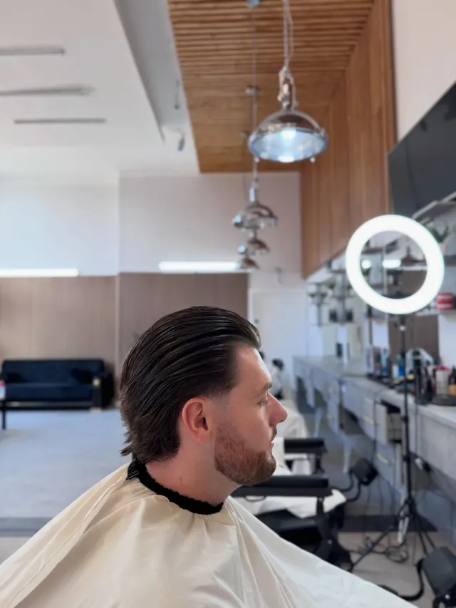 A long slicked-back cut in the chair, with the ring light and the row of stations behind